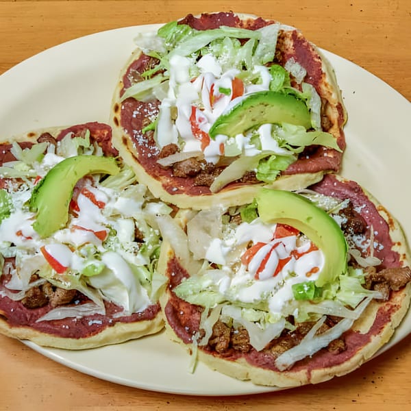 Three Latin-American sopes topped with refried beans, meat, lettuce, cheese, sour cream, and avocado slices.
