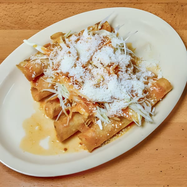 Golden fried taquitos topped with shredded cabbage, sauce, and crumbled white cheese on a white plate.