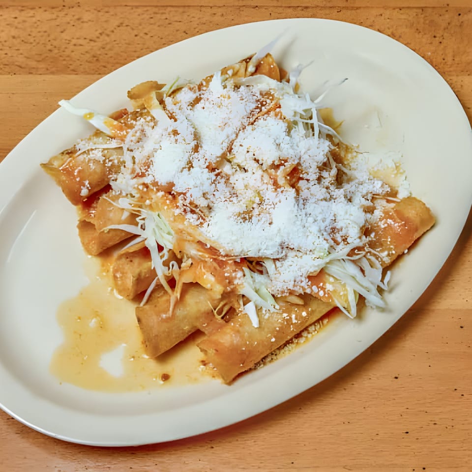 Golden fried taquitos topped with shredded cabbage, sauce, and crumbled white cheese on a white plate.